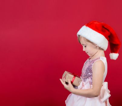 Dreaming funny cacusian cute child girl in santa hat looking at gift box celebrating happy 2021 New Year isolated on red background. Merry Christmas presents shopping sale.