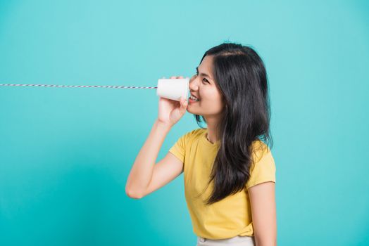 Portrait happy Asian beautiful young woman smile white teeth standing wear yellow t-shirt, She holding paper can telephone for talking, studio shot on blue background with copy space for text