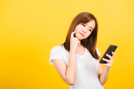 Asian happy portrait beautiful cute young woman teen standing wear t-shirt her using holding smart mobile phone and thinking looking to the phone isolated, studio shot yellow background with copy space