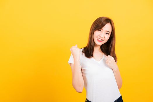 Asian happy portrait beautiful cute young woman teen standing wear t-shirt makes raised fists up celebrating her success looking to camera isolated, studio shot on yellow background with copy space