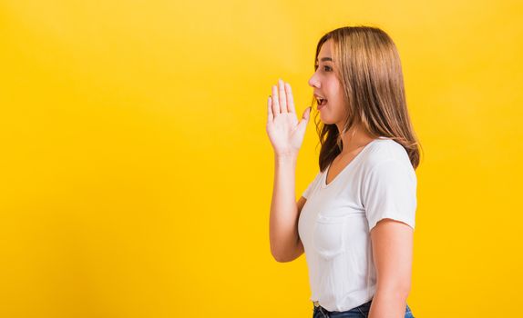 Asian Thai happy portrait beautiful young woman standing wear t-shirt hold a hand on mouth talking whispering secret rumor looking to camera isolated, studio shot on yellow background with copy space