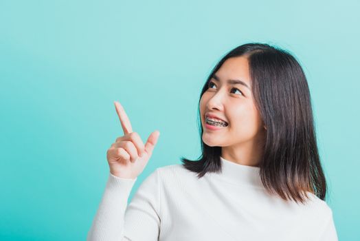 Portrait of Asian teen beautiful young woman smile have dental braces on teeth laughing point finger side away blank copy space, studio shot isolated on blue background, medicine and dentistry concept