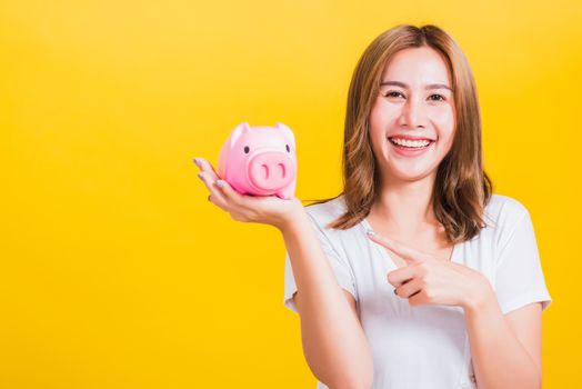 Portrait Asian Thai beautiful happy young woman smiling holding pink piggy bank with a lot money looking to camera and she point a finger to piggybank, isolated on yellow background, with copy space