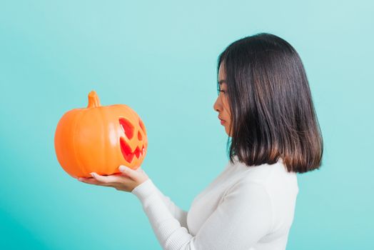 Portrait of Asian beautiful young woman holding orange model pumpkins and looking it, funny happy female with ghost pumpkins, studio shot isolated on blue background