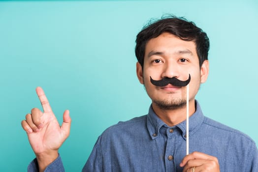 Portrait Asian happy handsome man posing he holding a funny mustache card or vintage fake moustaches on his mouth, studio shot isolated on blue background, Fathers day, November day concept