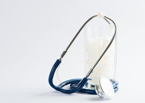 A glass full of white sugar cube sweet food ingredient and doctor stethoscope, studio shot isolated white background, health high blood risk of diabetes and calorie intake concept and unhealthy drink