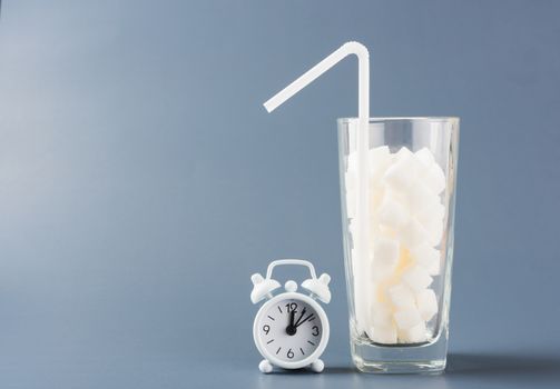 A glass full of white sugar cube sweet food ingredient and alarm clock, studio shot isolated on a gray background, health high blood risk of diabetes and calorie intake concept and unhealthy drink