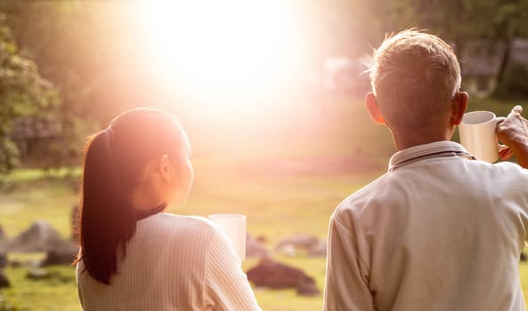 Rear view of granddaughter and grandfather drinking hot coffee outdoor and enjoying landscape during holiday on the mountain with sunlight in morning.