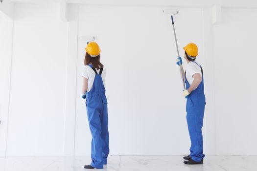 Two young workers in uniform of coverall and hardhat painting white wall