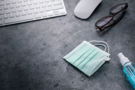 disposable surgical face mask and alcohol spray in working space with computer keyboard, mouse and eyeglasses, concept of flu prevention during situation of COVID-19 to stop pandemic, selective focus