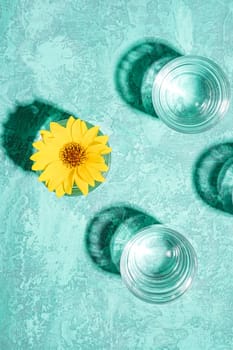 Fresh clear water drink with yellow flower in glass on turquoise background, hard light creative composition, top view