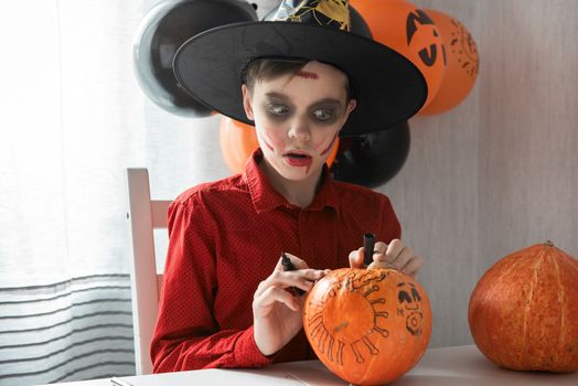 Teen boy in costume drawing coronavirus image on the pumpkin for the Halloween celebration. Halloween carnival with new reality with pandemic concept.