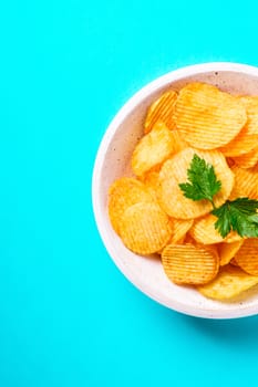 Fried corrugated golden potato chips with parsley leaf in wooden bowl on turquoise background, top view