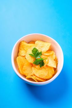 Fried corrugated golden potato chips with parsley leaf in ceramic bowl on blue background, angle view