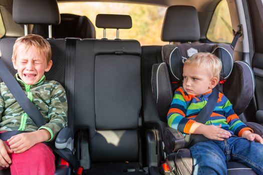 Two little boys sitting on a car seat and a booster seat buckled up upset and crying in the car. Children's Car Seat Safety