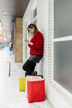 Adult woman looking at her cell phone in a mall with shopping bags on the floor