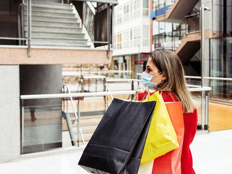 Adult woman walking through a mall with a face mask and carrying colorful shopping bags. Copy space.