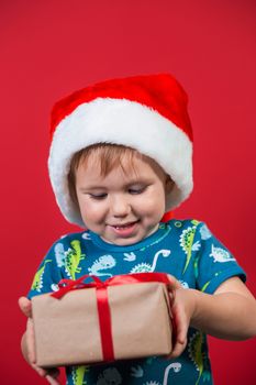 A little boy wearing a Santa hat tries to unpack a New Year's gift with enthusiasm and excitement on a red background