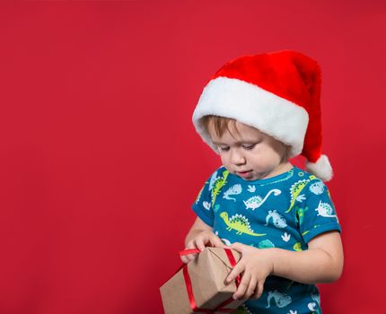 A little boy wearing a Santa hat tries to unpack a New Year's gift with enthusiasm and excitement on a red background