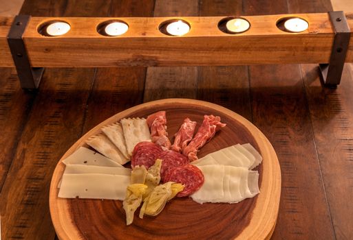 Charcuterie board on rustic wood with candles behind a spread of prosciutto, mozzarella cheese, Genoa salami, Fontina cheese and artisanal crackers.