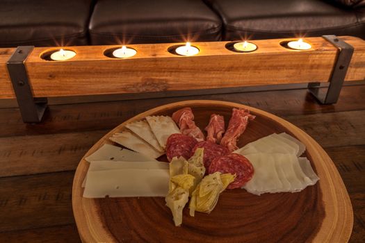 Charcuterie board on rustic wood with candles behind a spread of prosciutto, mozzarella cheese, Genoa salami, Fontina cheese and artisanal crackers.