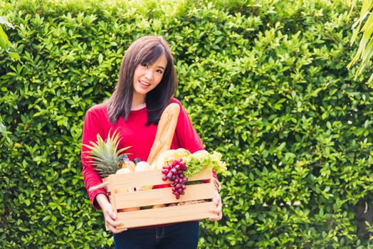 Portrait of Asian beautiful young woman farmer standing she smile and holding full fresh food raw vegetables fruit in a wood box in her hands on green leaves background