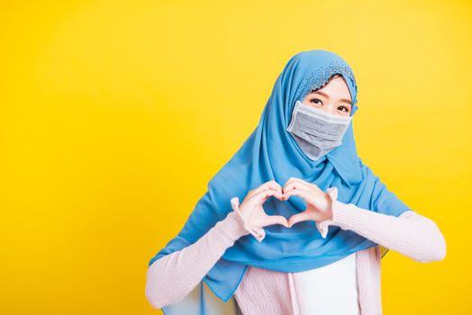 Asian Muslim Arab, Portrait of happy beautiful young woman Islam religious wear veil hijab and face mask protect quarantines disease coronavirus show hands finger heart figure isolated yellow