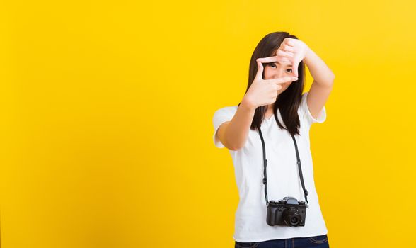 Portrait of happy Asian beautiful young woman photographer execute by hanging digital mirrorless photo camera on neck and show hand sign mark frame for photography, isolated on yellow background