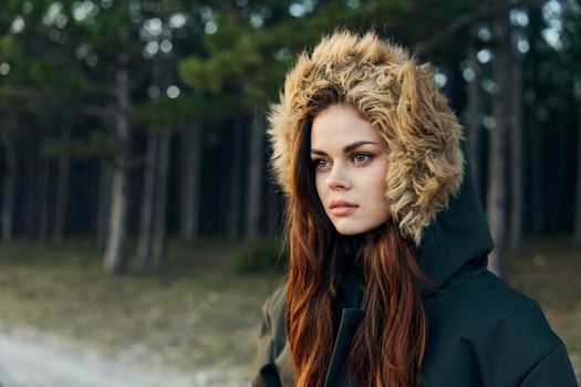 A woman in a warm autumn jacket with a hood looks to the side on nature against the background of trees. High quality photo