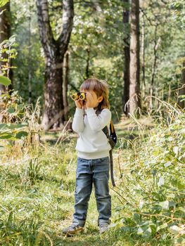 Curious boy is hiking in forest. Outdoor leisure activity for kids. Child looks through binoculars on tree foliage. Sunny day at autumn or summer day.