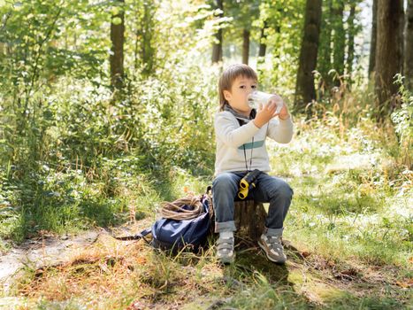 Little explorer on hike in forest. Boy with binoculars sits on stump and drinks water from bottle. Outdoor leisure activity for children. Summer journey for young tourist.