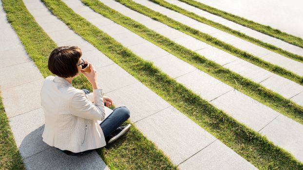 Woman with short haircut and eyeglasses sits in park with take away cup of coffee. Woman enjoys summer sun. Casual clothes, urban lifestyle of millennials.