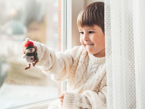 Kid with felt decorative snowman for Christmas tree. Boy in cable-knit oversized sweater. Cozy outfit for snuggle weather. Funny character with red Santa Claus hat. Winter holiday spirit. New year.