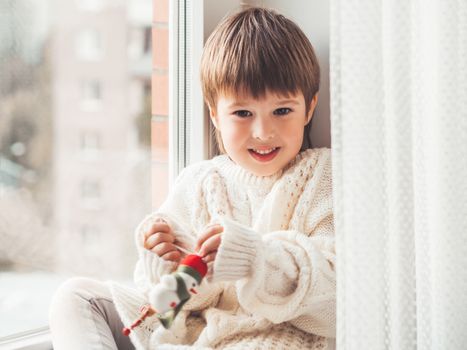 Kid with felt decorative snowman for Christmas tree. Boy in cable-knit oversized sweater. Cozy outfit for snuggle weather. Funny character with red Santa Claus hat. Winter holiday spirit. New year.