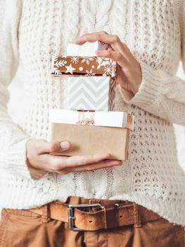 Woman in cable-knit white sweater with Scandinavian pattern and brown chinos trousers with leather belt. Stack of Christmas presents in hands. Casual outfit for New Year celebration.