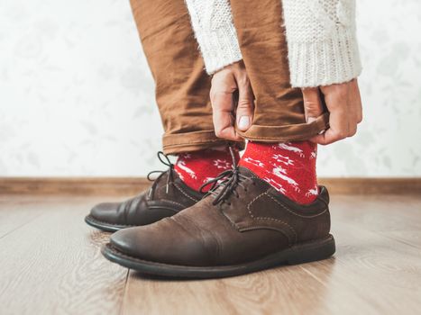 Young man pulls up leg of his chinos trousers to show bright red socks with reindeers on them. Scandinavian pattern. Winter holiday spirit. Casual outfit for New Year and Christmas celebration.