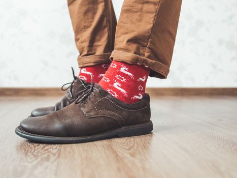 Young man pulls up leg of his chinos trousers to show bright red socks with reindeers on them. Scandinavian pattern. Winter holiday spirit. Casual outfit for New Year and Christmas celebration.