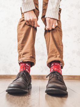 Young man pulls up leg of his chinos trousers to show bright red socks with reindeers on them. Scandinavian pattern. Winter holiday spirit. Casual outfit for New Year and Christmas celebration.