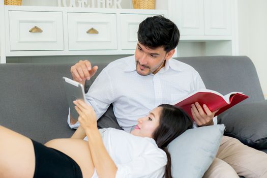 Asian couple family young mother and husband using a tablet at home, woman and man is pregnant lying on sofa looking tablet read book.