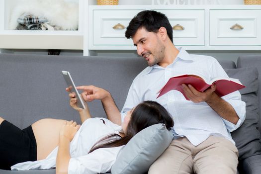 Asian couple family young mother and husband using a tablet at home, woman and man is pregnant lying on sofa looking tablet read book.