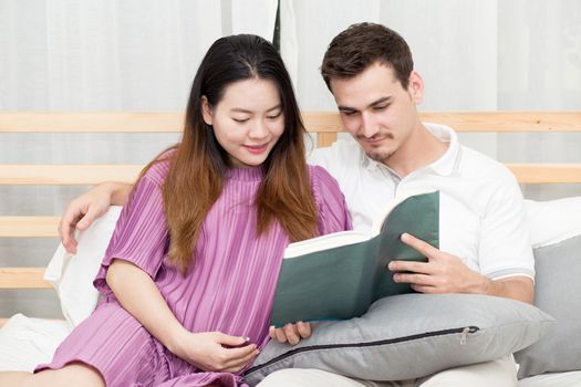 Asian couple family young mother and husband reading book at home, woman and man is pregnant lying on sofa and relax.