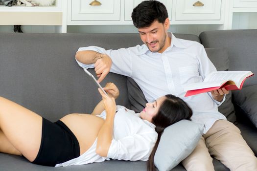 Asian couple family young mother and husband using a tablet at home, woman and man is pregnant lying on sofa looking tablet read book.