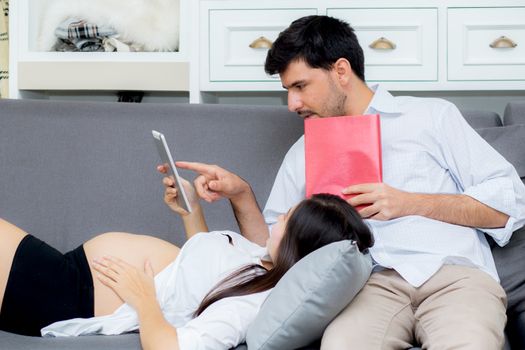 Asian couple family young mother and husband using a tablet at home, woman and man is pregnant lying on sofa looking tablet read book.