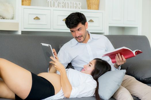 Asian couple family young mother and husband using a tablet at home, woman and man is pregnant lying on sofa looking tablet read book.