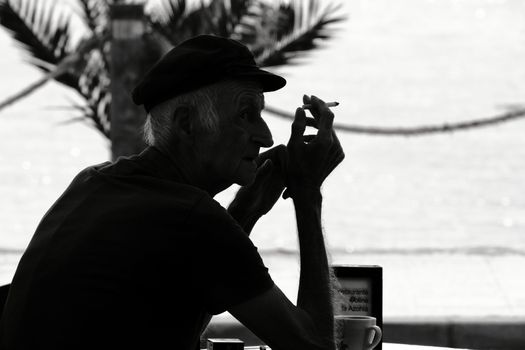 Alicante, Spain- October 1, 2019: Old sailor smoking alone on the terrace of a bar on the beach in Spain. Monochrome picture.