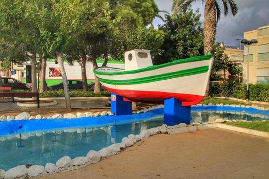 Isla Plana, Murcia, Spain- October 7, 2019: Colorful fishing boat surrounded by vegetation and a fountain in the main square of Isla Plana village in Cartagena province, Murcia, Spain