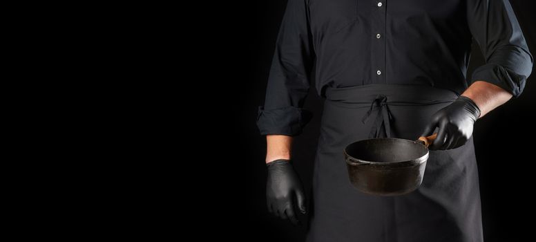 cook in black uniform and latex gloves holds an empty round vintage black cast iron pan in front of him, copy space