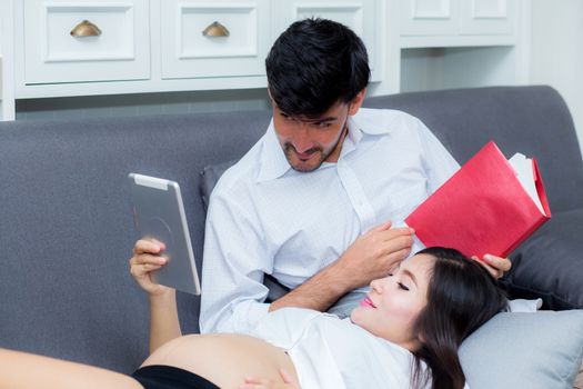 Asian couple family young mother and husband using a tablet at home, woman and man is pregnant lying on sofa looking tablet read book.