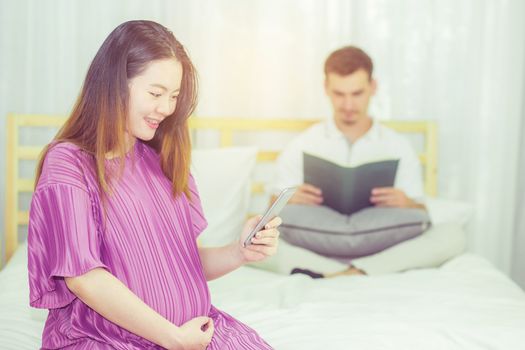 Asian pregnant people and expectation concept - happy pregnant woman holding mobile phone sitting on bed and touching belly at home with father reading book background.