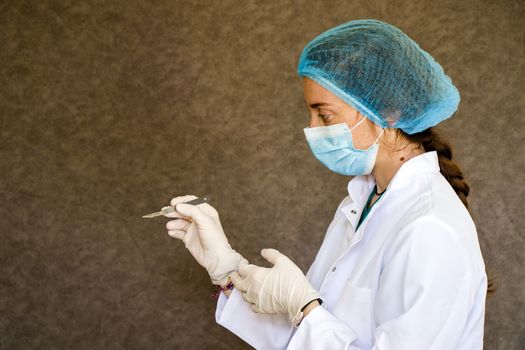 Doctors portrait with mask, glove and uniform and surgery blade in hand on the gray background.